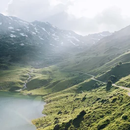 Wanderweg entlang eines Bergsees mit grünen Hängen und schneebedeckten Gipfeln