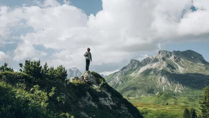 Mann steht auf Berggipfel mit Blick auf bewachsene Berge und bewölkten Himmel