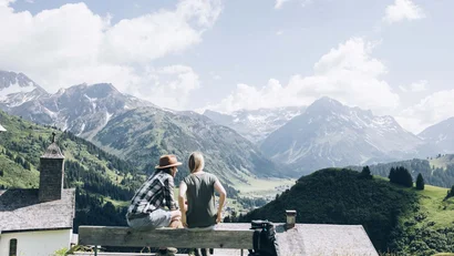Zwei Personen sitzen auf einer Bank mit Blick auf Alpen und Dorf