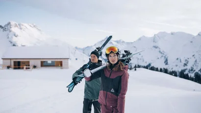 Zwei Skifahrer tragen Skier vor schneebedeckten Bergen und einer Hütte