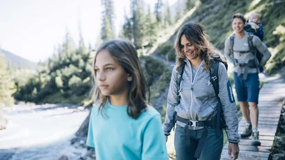 Familie wandert auf Holzsteg neben Fluss in bergiger Waldlandschaft