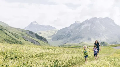 Kinder und Frau laufen fröhlich durch blühende Almwiese vor Bergkulisse