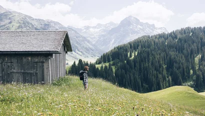 Wanderer steht neben Hütte auf blühender Bergwiese mit Alpen im Hintergrund