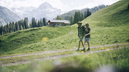 Paar wandert auf einem Weg in einer grünen Berglandschaft mit einer Hütte im Hintergrund