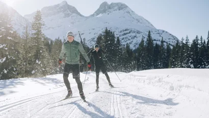 Zwei Langläufer auf verschneiter Strecke mit Bergen und Tannen im Hintergrund